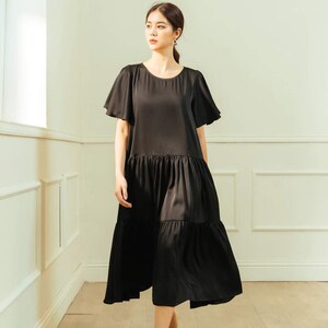 May include: A woman wearing a black tiered midi dress with short, flared sleeves. She is walking on a wooden floor in a room with white walls.