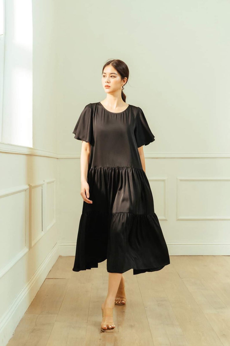 May include: A woman wearing a black tiered midi dress with short, flared sleeves. She is walking on a wooden floor in a room with white walls.