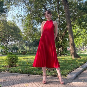 Pode incluir: Uma mulher vestindo um vestido midi vermelho plissado com decote halter. Ela est&aacute; em p&eacute; em um caminho de paralelep&iacute;pedos em um parque.