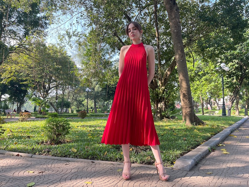 Pode incluir: Uma mulher vestindo um vestido midi vermelho plissado com decote halter. Ela est&aacute; em p&eacute; em um caminho de paralelep&iacute;pedos em um parque.