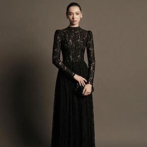 May include: A long, black lace dress with long sleeves and a high neckline. The dress has a fitted bodice and a flowing skirt. A small black clutch is held in the hand. The dress is suitable for formal events.