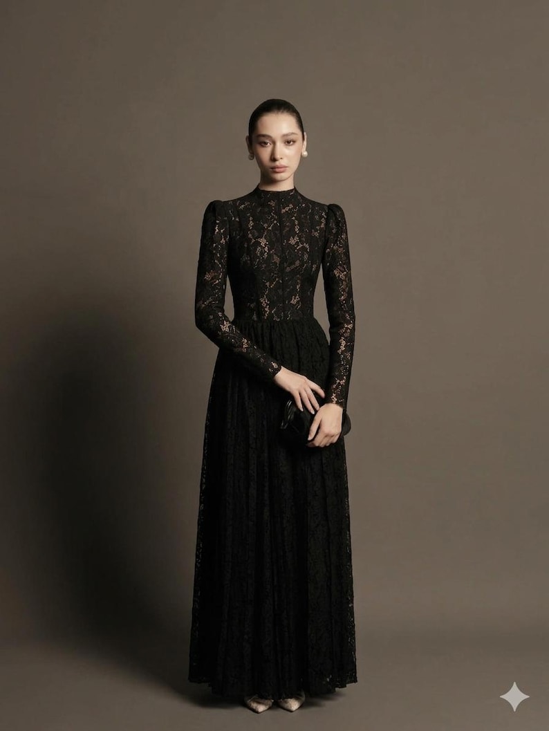 May include: A long, black lace dress with long sleeves and a high neckline. The dress has a fitted bodice and a flowing skirt. A small black clutch is held in the hand. The dress is suitable for formal events.