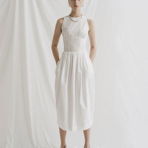 May include: A white sleeveless dress with a fitted bodice and a gathered skirt. The dress has a scoop neckline and a silver chain necklace. The model is wearing black lace-up shoes.