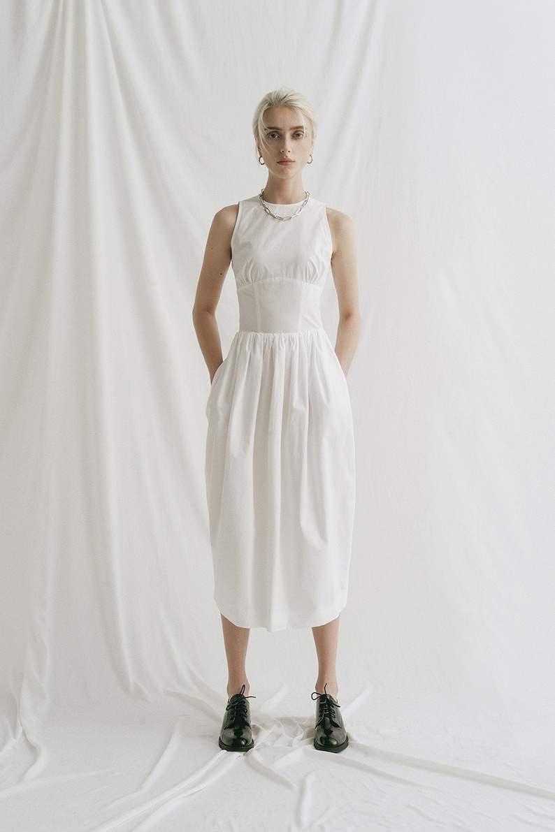 May include: A white sleeveless dress with a fitted bodice and a gathered skirt. The dress has a scoop neckline and a silver chain necklace. The model is wearing black lace-up shoes.