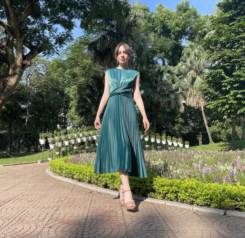 Peut inclure: Une femme portant une robe midi pliss&eacute;e verte avec un corsage crois&eacute; et une finition satin&eacute;e. Elle se tient dans un jardin avec de la verdure et des fleurs.