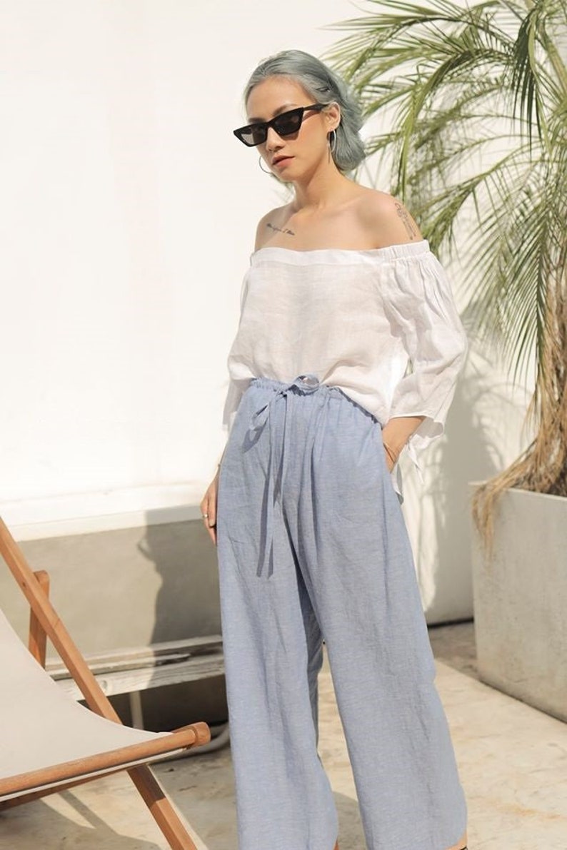 Puede incluir: Una mujer con un top blanco de hombros descubiertos y pantalones anchos de color azul claro. Lleva gafas de sol negras y tiene el pelo gris.