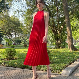 May include: A woman wearing a red pleated midi dress with a halter neckline. She is walking on a brick pathway in a park.
