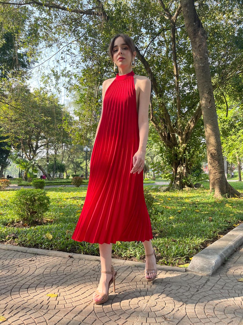 May include: A woman wearing a red pleated midi dress with a halter neckline. She is walking on a brick pathway in a park.