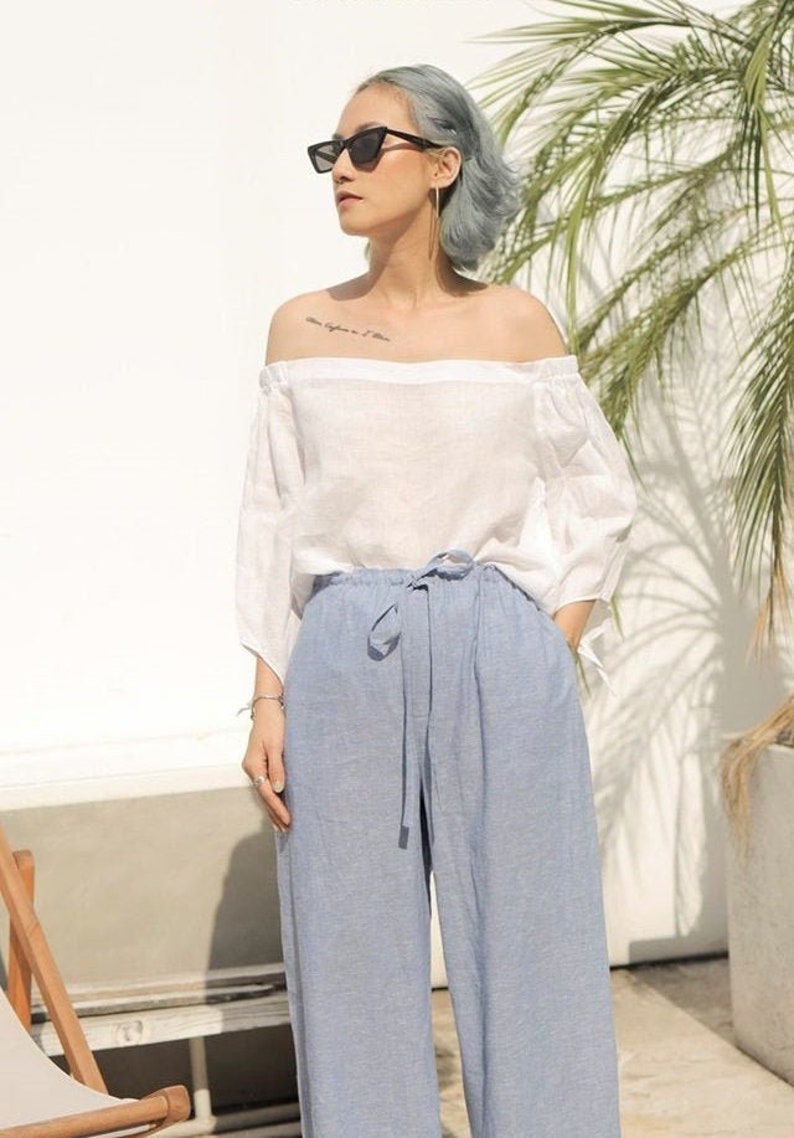 Puede incluir: Una mujer con una blusa blanca de hombros descubiertos y pantalones anchos de color azul claro. Lleva gafas de sol negras y tiene el pelo corto y gris.