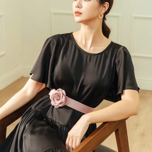 May include: A woman wearing a black dress with a pink flower accent on the belt. The dress has a ruffled skirt and short sleeves. She is sitting on a wooden chair.