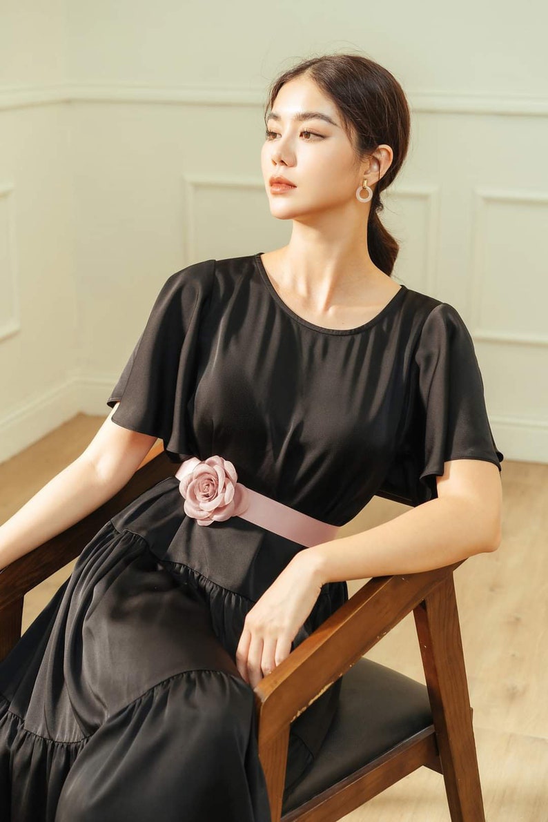 May include: A woman wearing a black dress with a pink flower accent on the belt. The dress has a ruffled skirt and short sleeves. She is sitting on a wooden chair.
