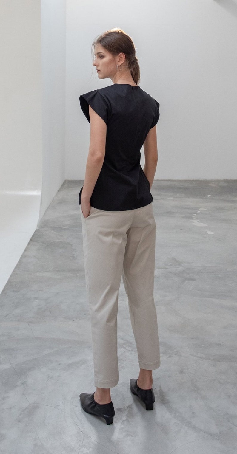 K&ouml;nnte beinhalten: Eine Frau tr&auml;gt ein schwarzes, kurz&auml;rmeliges Oberteil mit Rundhalsausschnitt und beigefarbene Hosen. Sie tr&auml;gt schwarze Schuhe.