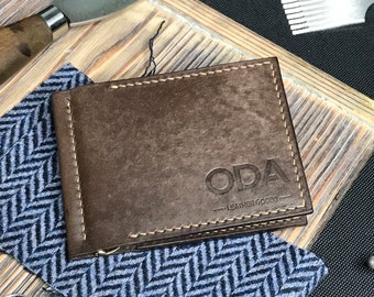 May include: A brown leather wallet with the text "ODA Leather Goods" embossed on the front. The wallet is sitting on a blue and white patterned fabric.