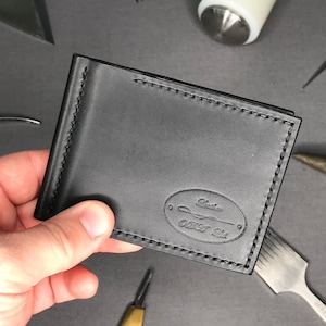Peut inclure: Portefeuille en cuir noir avec un porte-billets et un bord cousu. Le portefeuille a un logo ovale avec le texte "Leather Craft Co."