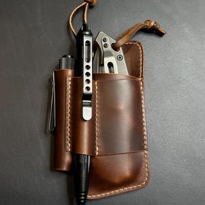 May include: A brown leather pocket organizer with a pen, knife, and a pen. The organizer has a stitched border and a leather strap. The pen is black and silver, and the knife is silver.