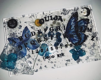 Peut inclure: Un plateau de Ouija blanc avec des lettres et des chiffres noirs, décoré de papillons bleus et de fleurs pressées. Le plateau a un croissant de lune dans le coin supérieur droit et un planchette en forme de cœur avec une fleur bleue.