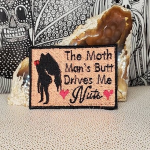 May include: A brown embroidered patch with black text that reads "The Moth Man's Butt Drives Me Nuts". The patch features a black silhouette of a person with a red heart above their head. Two pink hearts are on either side of the text.
