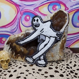 Könnte beinhalten: Weißer gestickter Aufnäher mit einer Cartoon-Figur mit großem Kopf und langen Gliedmaßen, die auf einem braunen Felsen steht. Der Aufnäher befindet sich auf einer weißen Häkelspitze.