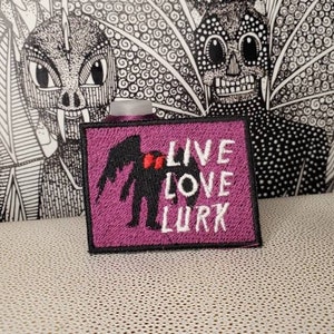 Op de afbeelding: Paarse geborduurde patch met een zwart silhouet van een beest met vleugels en rode harten. De tekst "LIVE LOVE LURK" is geborduurd in wit.