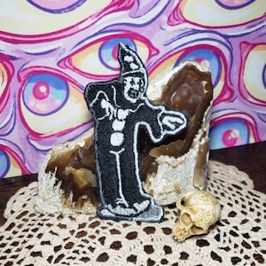 May include: Black and white embroidered patch of a clown with a pointed hat, standing on a rock with a skull beside it. The background is a pink and purple pattern of eyes.