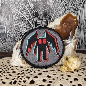 May include: A black and gray embroidered patch with a red and blue creature with wings. The creature has two large black eyes. The patch is on a brown rock and a white doily.