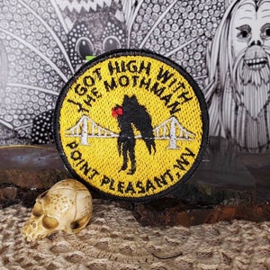 May include: Yellow embroidered patch with black text that reads "I got high with the Mothman Point Pleasant WV". The patch features a black silhouette of the Mothman with red eyes and a bridge in the background.
