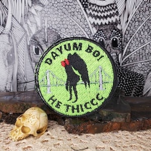 Può includere: Toppa ricamata verde e nera con il testo "Dayum Boi He Thicc" e una silhouette di una coppia che si bacia davanti a un ponte.