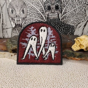 Może przedstawiać: Naszywka haftowana w kolorze bordowym z trzema białymi duchami w leśnym otoczeniu. Duchy nie mają twarzy i są przedstawione z długimi nogami i ramionami.