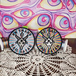 May include: Two embroidered patches with a rainbow background and a black goat head with horns and a pentagram. The text on the patches reads "Be Trans Hail Satan" and "Be Gay Hail Satan".