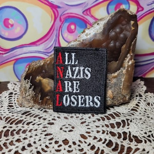 May include: A black square patch with the text "ALL NAZIS ARE LOSERS" in white and red lettering. The patch is displayed on a brown and beige geode, set against a colorful, psychedelic background.