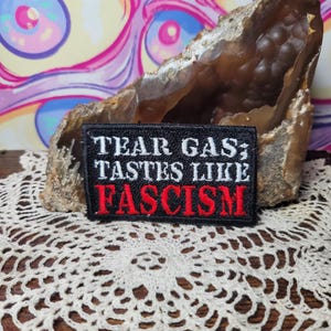 May include: A black patch with white text reading "TEAR GAS TASTES LIKE FASCISM" in a red font. The patch is set against a brown geode and a cream-colored lace doily, with a colorful, psychedelic background.