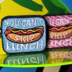 May include: Two embroidered patches featuring a hot dog and the text "You can't skip lunch". The patch on the left is blue with white text. The patch on the right is pink with white text.
