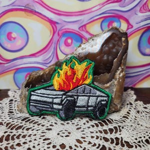 May include: Embroidered patch featuring a gray car engulfed in flames with a green border. The flames are depicted in shades of red, orange, and yellow. The patch is placed on a geode and a white lace doily.