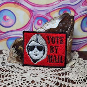 May include: A square, red patch with the words "VOTE BY MAIL" in black, and a white illustration of a person wearing sunglasses and a hood. The patch is embroidered with black thread. The patch is displayed on a decorative rock.