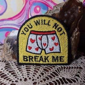 Puede incluir: Parche bordado con el texto "YOU WILL NOT BREAK ME" sobre una imagen de dibujos animados de ropa interior blanca con detalles de corazones rojos. El parche tiene un fondo amarillo y un borde negro.