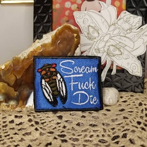 May include: A blue embroidered patch featuring a cicada insect illustration and the words "Scream Fuck Die" in white script. The patch has a black border and is set against a neutral background.