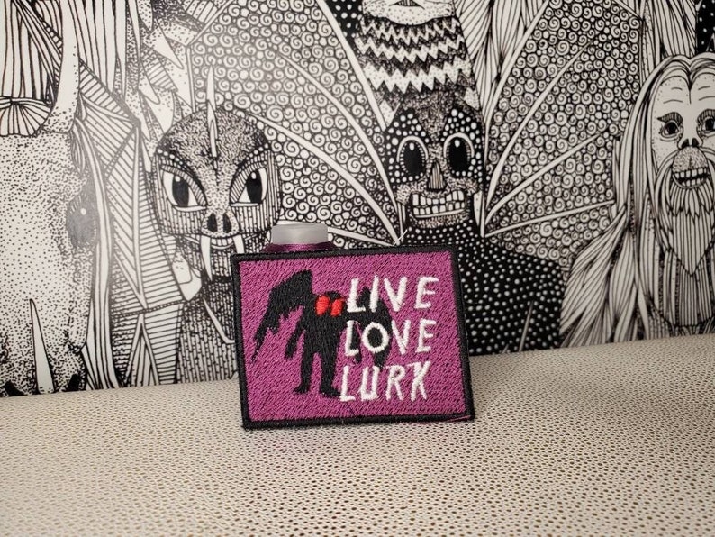 May include: Purple embroidered patch with a black silhouette of a creature with wings and red hearts. The text "LIVE LOVE LURK" is embroidered in white.