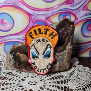 Op de afbeelding: Geborduurde patch met de tekst "FILTH IS MY POLITICS" in zwart en oranje. De patch heeft een gestileerd gezicht met blauwe ogen en een rode omtrek. De patch is geplaatst tegen een achtergrond van een geode en een patroonachtergrond.