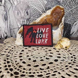 May include: Red embroidered patch with a black silhouette of a creature with the text "Live Love Lurk" in black.