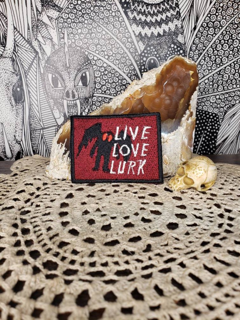May include: Red embroidered patch with a black silhouette of a creature with the text "Live Love Lurk" in black.