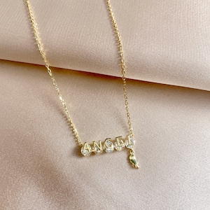 Pu&ograve; includere: Collana con catena dorata con un ciondolo che scrive "ANGEL" con piccole pietre trasparenti su ogni lettera e un piccolo ciondolo ad ala dorata.