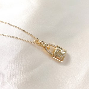 May include: A gold-colored necklace featuring a padlock pendant. The pendant is adorned with small, clear gemstones and a crescent moon and star design. The necklace chain is delicate and thin, and the background is a soft, white fabric.