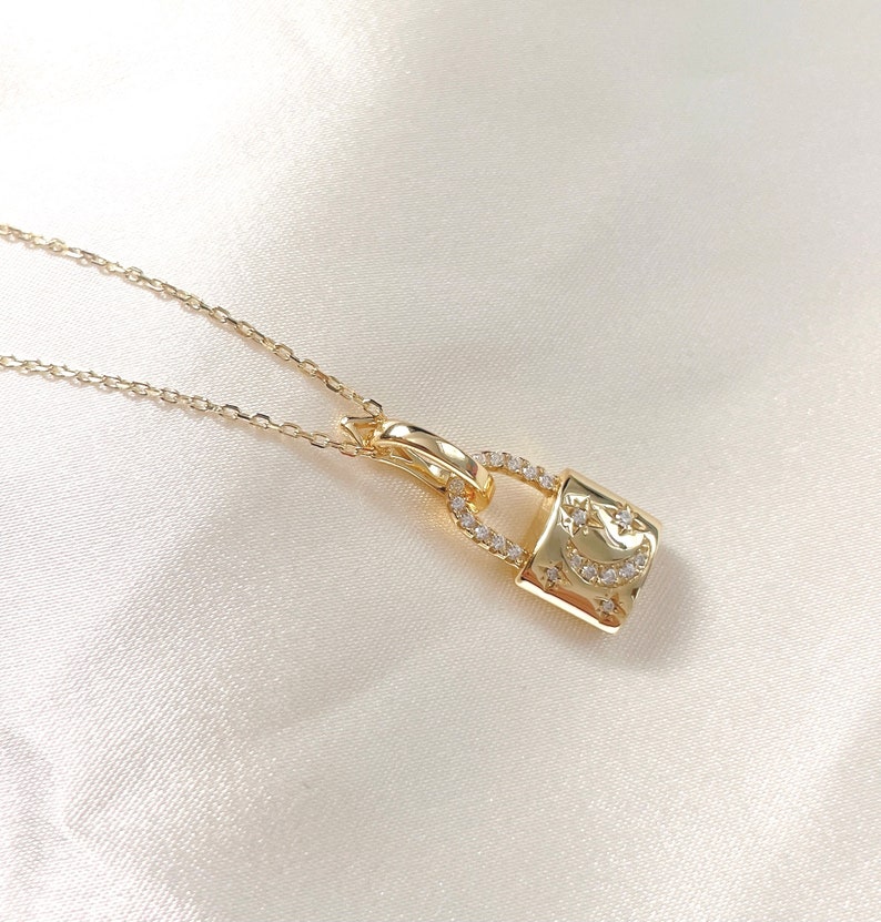 May include: A gold-colored necklace featuring a padlock pendant. The pendant is adorned with small, clear gemstones and a crescent moon and star design. The necklace chain is delicate and thin, and the background is a soft, white fabric.
