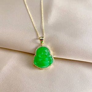Puede incluir: Un collar de cadena dorada con un colgante de jade verde que presenta una figura tallada de Buda riendo. El colgante está rodeado por una fila de piedras de circonita cúbica transparentes.