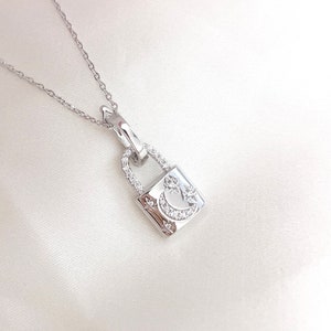 May include: A silver-colored necklace featuring a small, square pendant shaped like a padlock. The pendant is adorned with a crescent moon and star design, embellished with small, sparkling stones. The necklace hangs on a delicate chain.