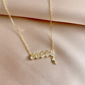 Pu&ograve; includere: Una collana con catena dorata con un ciondolo che dice "ANGEL" con un piccolo ciondolo a forma di ala appeso alla "L".