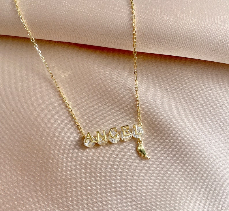 Pu&ograve; includere: Una collana con catena dorata con un ciondolo che dice "ANGEL" con un piccolo ciondolo a forma di ala appeso alla "L".