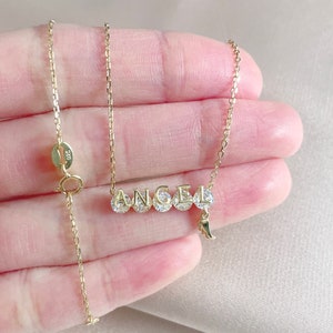 Pu&ograve; includere: Collana con catena dorata e ciondolo che compone la parola "ANGEL" in lettere maiuscole con pietre di zircone cubico trasparenti. Il ciondolo ha un piccolo ciondolo a forma di ala d'angelo che pende.