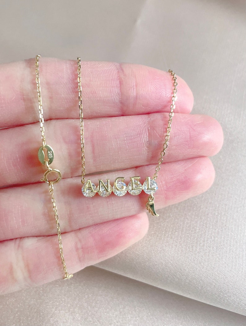 Pu&ograve; includere: Collana con catena dorata e ciondolo che compone la parola "ANGEL" in lettere maiuscole con pietre di zircone cubico trasparenti. Il ciondolo ha un piccolo ciondolo a forma di ala d'angelo che pende.
