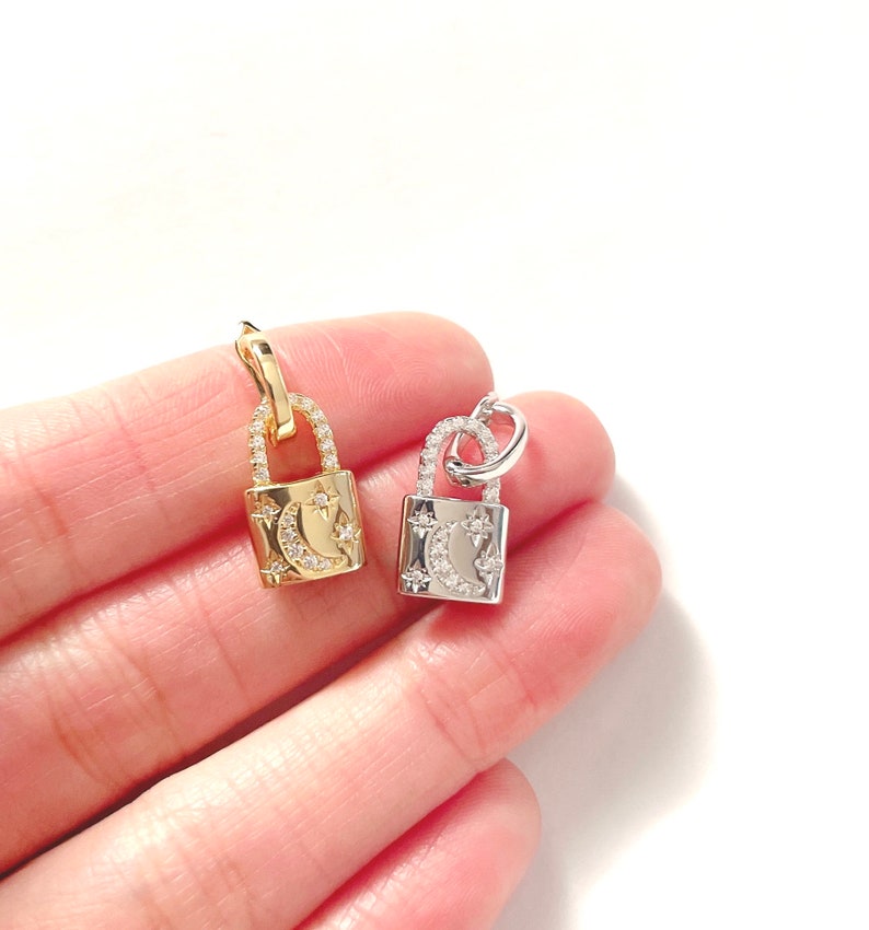 May include: Two small padlock pendants, one gold-toned and one silver-toned, each adorned with a crescent moon and star design. The pendants are embellished with small, sparkling crystals, adding a touch of elegance to the jewelry.